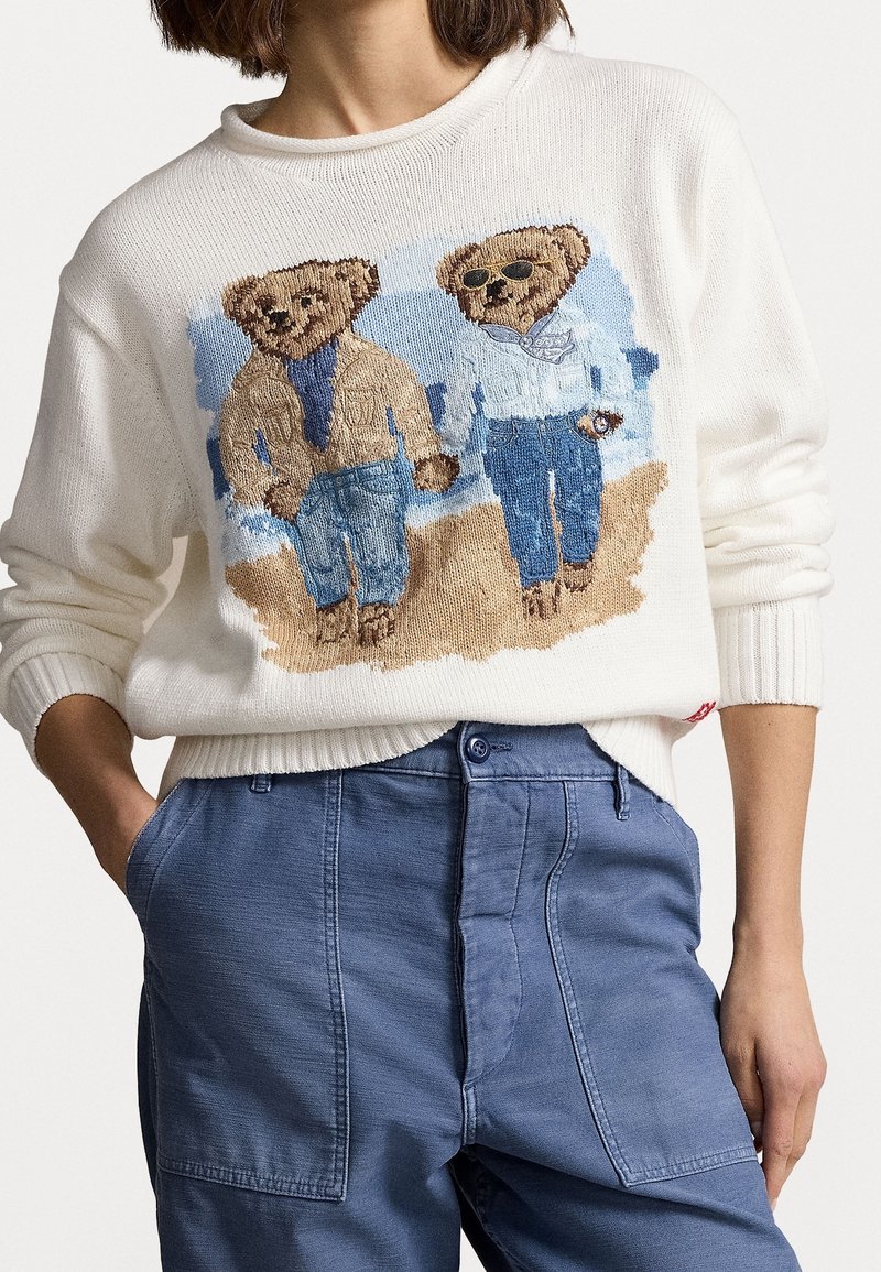 Bílý pletený svetr s motivem dvou medvědů v ležérním oblečení, jeden má sluneční brýle, na modrém prošitém pozadí. Kombinováno s modrými kalhotami.