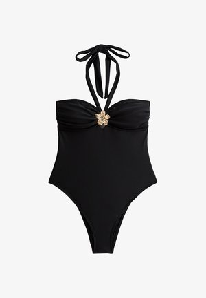 Bañador de una pieza negro con tirantes al cuello, fruncido en el busto y un acento de flor dorada en el centro. Textura suave y cobertura total.