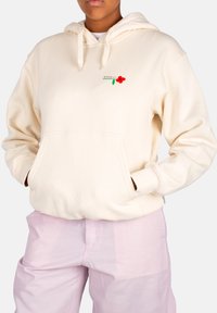 Cremefarbene Kapuzenjacke mit Fronttasche, Kordelzug-Kapuze und rotem Blumenstick. Kombiniert mit hellrosa Hosen. Baumwollmaterial, lockere Passform.