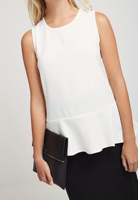Top blanco sin mangas con un acabado texturizado, que presenta una cintura con peplum, combinado con un bolso clutch negro, sostenido en una mano.
