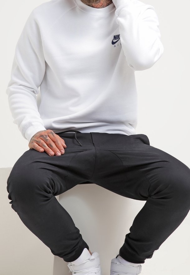 Witte Nike-sweatshirt met een ronde hals en een blauw logo, gecombineerd met zwarte taps toelopende joggers met voorzakken en een gladde textuur.