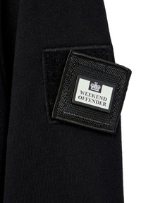 Sort stof med en tekstureret overflade har et kvadratisk patch, der viser "WEEKEND OFFENDER"-logoet i hvidt og sort med et kroneemblem.