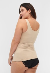 Frau mit langen braunen Haaren, die ein beiges Tanktop und schwarze Unterwäsche mit Spitze trägt, leicht seitlich zugewandt vor einem schlichten Hintergrund.