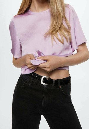 Frau trägt ein helllila kurzärmeliges Oberteil und schwarze Jeans mit einem schwarzen Gürtel, hält und hebt leicht den Saum des Shirts an.