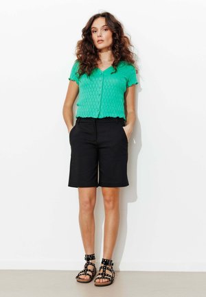 Femme aux cheveux bruns bouclés portant un haut vert à manches courtes boutonné, un short noir arrivant aux genoux, et des sandales noires cloutées, debout contre un mur blanc.