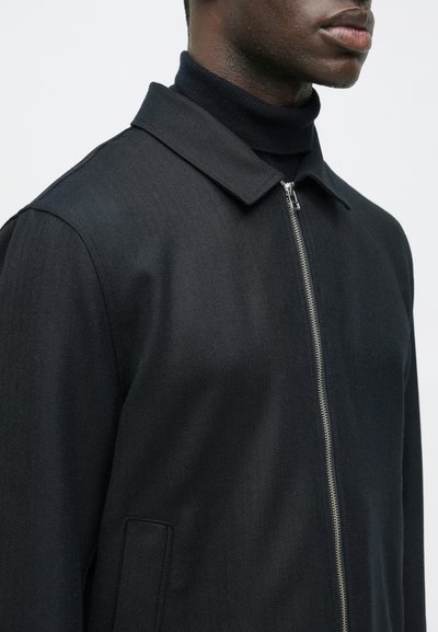 Vīrietis melnā ar rāvējslēdzēju aizveramā jaciņā virs melnas tārpa kreklā, redzams no kakla līdz vidus ķermenim uz viendabīgas fonā.