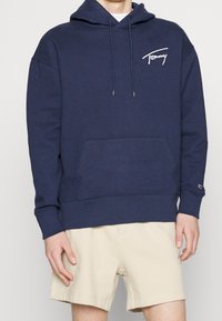 Mörkblå hoodie av bomullsblandning, med en framficka, justerbar dragsko på huvan och en vit logotyp på bröstet.