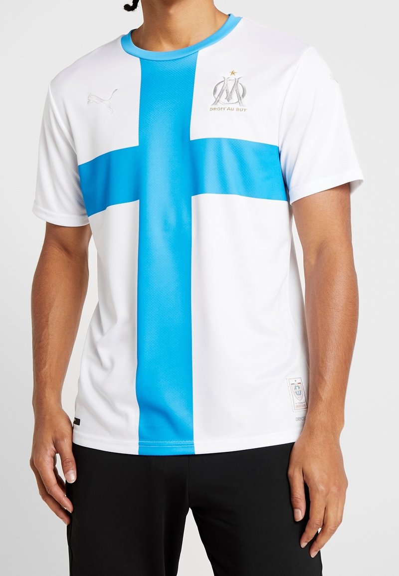 Valkoinen urheiluhaalari, jossa on voimakas sininen risti -kuvio, logo brodeerattu rintaan, lyhyet hihat ja pyöreä kaula-aukko.