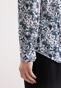 Manica di camicia bianca con motivo floreale blu, polsino abbottonato e una mano posata accanto a pantaloni neri.