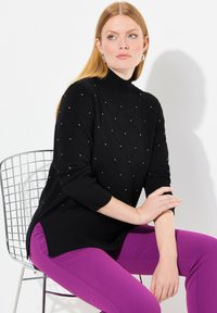 Svart polokrage tröja med ribbat mönster och diamantmönstrade utsmyckningar. I kombination med lila byxor. Modell som sitter på en trådstol.