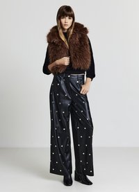 Braune ärmellose Weste aus Kunstpelz über einem schwarzen Oberteil, kombiniert mit weiten schwarzen Lederhosen mit silbernen Punktemustern. Schwarze Stiefel mit Absatz.