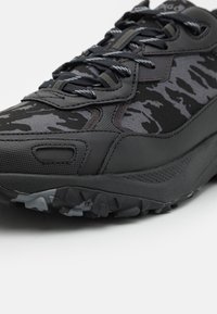 Chaussure de sport noire avec une tige texturée présentant un motif camouflage en nuances de gris. Semelle extérieure en caoutchouc durable avec des crampons d'adhérence.