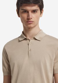 Beige pikétröja med klassisk krage, korta ärmar och treknappsstängning, tillverkad av mjukt, texturerat tyg.