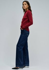 Mujer de pie con las manos en los bolsillos, vestida con una blusa roja de manga larga, vaqueros de pierna ancha de color azul oscuro y tacones altos negros de punta.
