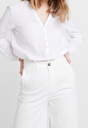 Personne portant une chemise blanche à manches longues boutonnée, rentrée dans un pantalon blanc taille haute à jambes larges, les mains posées sur les hanches.