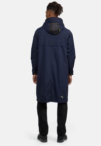Námořnická modrá dlouhá bunda do deště s kapucí, která má kontrastní černou podšívku kapuce a logo nášivku na dolní části zad. Lehce a elegantní design.