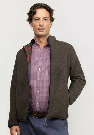 Hombre vistiendo una chaqueta de forro polar verde oscuro sobre una camisa rosa a cuadros con pantalones azules, de pie frente a un fondo claro y liso.