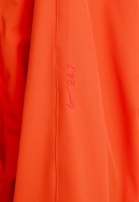 Nike-jakke med en lys oransje stoff, glatt tekstur, med en rosa Nike-logo og "24.7" trykket på ermet.