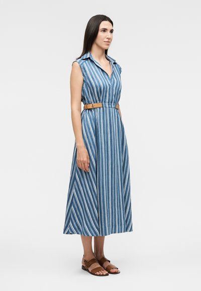 Γυναίκα που φοράει μπλε-λευκό ριγέ μίντι φόρεμα χωρίς μανίκια με καφέ ζώνη και καφέ σανδάλια ανοιχτά μπροστά, σε μονόχρωμο φόντο.