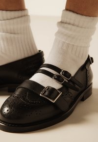 Μαύρα δερμάτινα παπούτσια Mary Jane με τρεις λουράκια με αγκράφες, φορεμένα με λευκές ριμππέρ κάλτσες σε ανοιχτόχρωμο δέρμα.