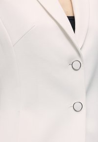 Λευκό σακάκι με βαθύ V-λαιμόκοψη, δύο μεγάλα στρογγυλά ασημένια κουμπιά και μαύρο εσώρουχο που φαίνεται από κάτω.
