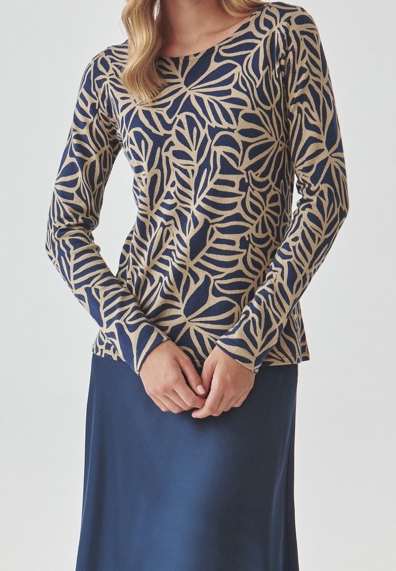 Femme portant un haut beige à manches longues avec un motif de feuilles bleu marine, associé à une jupe unie bleu marine, les mains jointes délicatement devant.