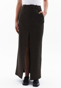 Larga falda marrón con una abertura frontal, hecha de tela texturada. Presenta bolsillos laterales y una cintura ajustada. Combinada con zapatos negros.