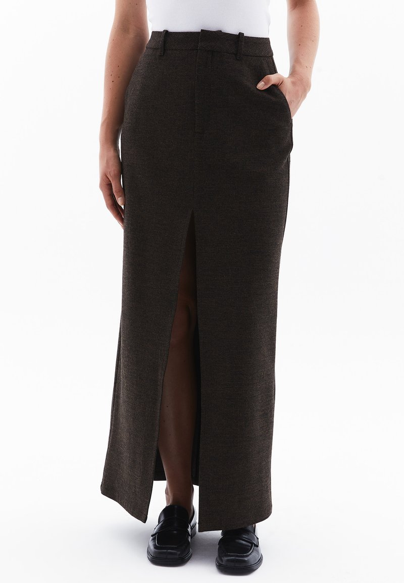 Larga falda marrón con una abertura frontal, hecha de tela texturada. Presenta bolsillos laterales y una cintura ajustada. Combinada con zapatos negros.