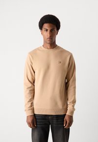 Sweat-shirt beige clair avec des poignets et un ourlet côtelés, arborant une petite étiquette logo sur la poitrine, porté avec un jean noir. Texture douce et lisse.