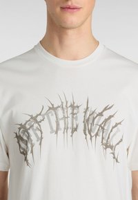 Hvid bomulds t-shirt med et grafisk print, der står "OFF THE WALL" i sølv med tornlignende accenter, korte ærmer og rund halsudskæring.