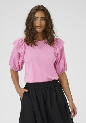 Hullámos barna hajú nő rózsaszín, fodros ujjú, csipkedíszes felsőt és magas derekú fekete szoknyát visel, egyik kezét a zsebében tartva pózol.