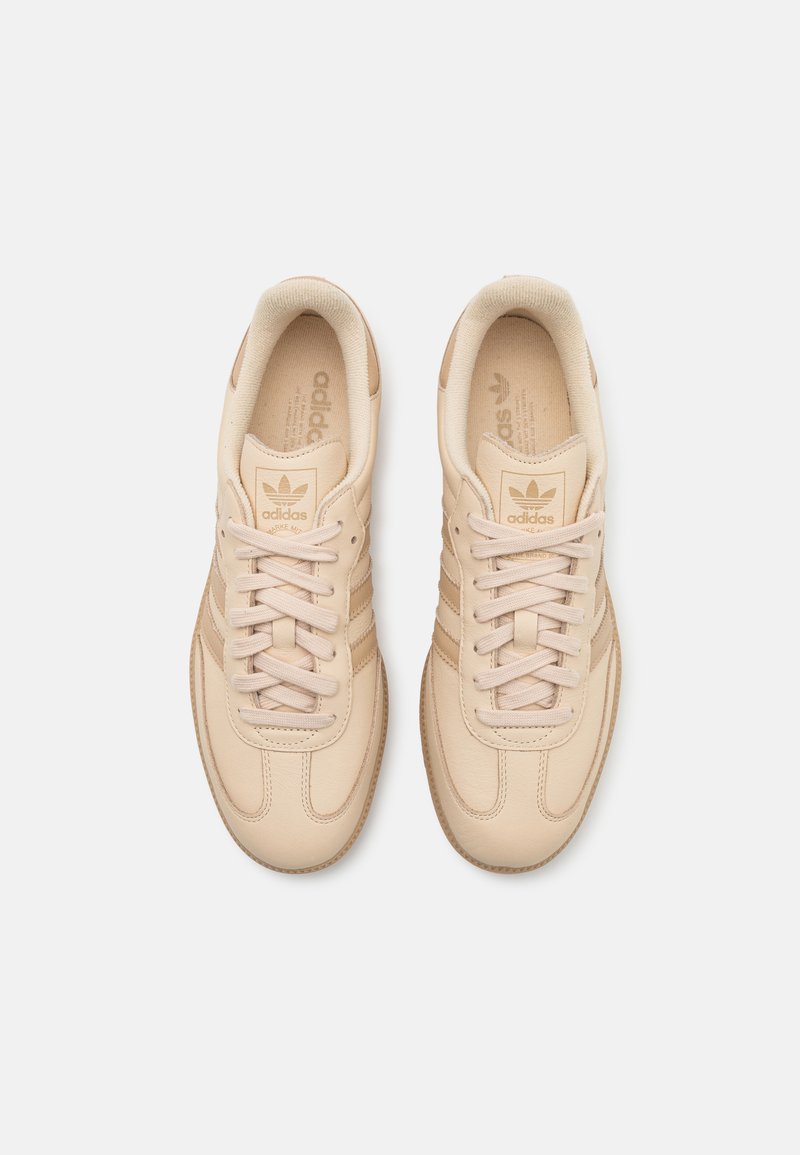 Zapatillas de cuero beige con una textura suave, que cuentan con cordones tonales, tres rayas diagonales y un logo grabado en la lengüeta.