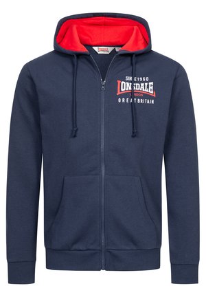 Granatowa bluza z kapturem na zamek z czerwonym wnętrzem, kapturem na sznurku, przednimi kieszeniami oraz białym napisem logo na piersi. Tkanina z mieszanki bawełny, swobodny krój.
