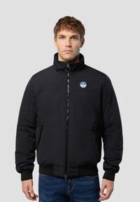 Schwarze Pufferjacke mit hohem Kragen, Frontreißverschluss, seitlichen Taschen und einem runden Logo auf der Brust. Glatte Textur und taillierte Passform.