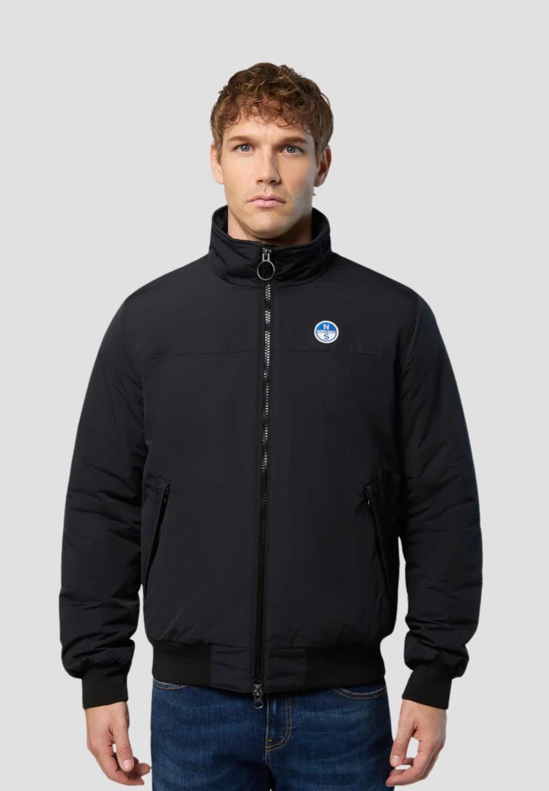 Schwarze Pufferjacke mit hohem Kragen, Frontreißverschluss, seitlichen Taschen und einem runden Logo auf der Brust. Glatte Textur und taillierte Passform.