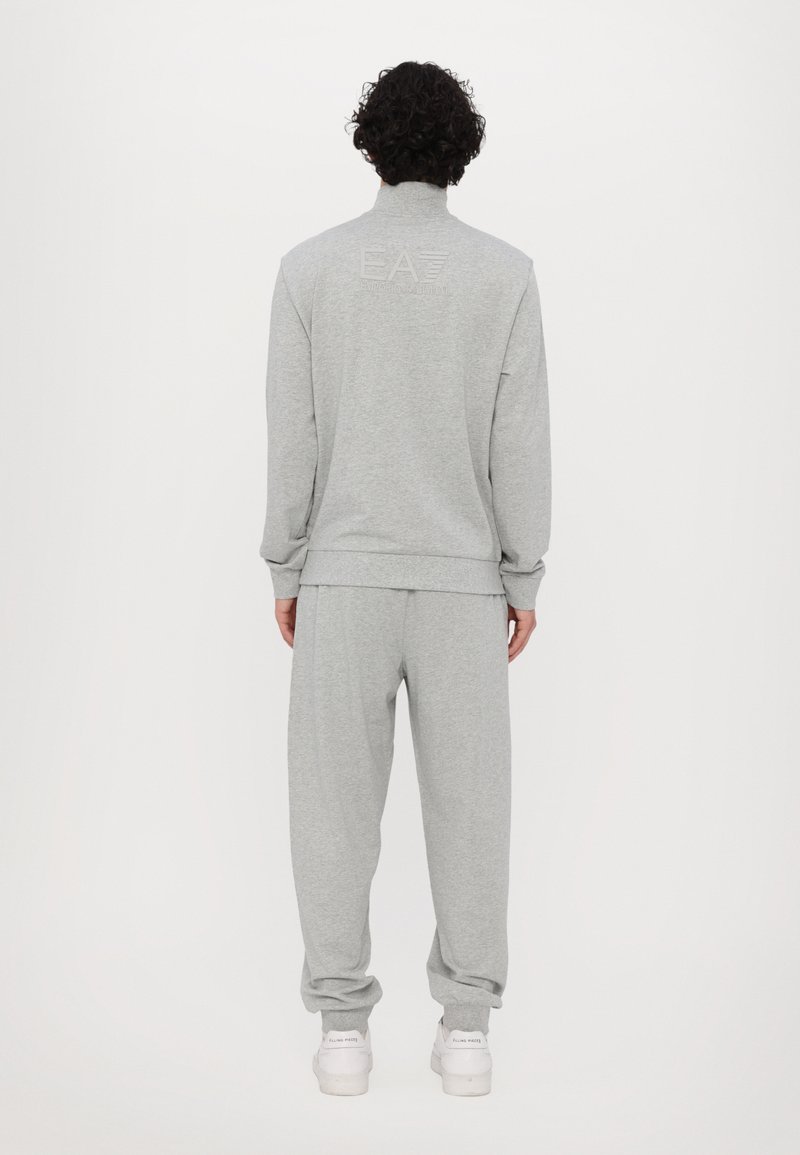 Survêtement gris composé d'un haut zippé avec un col haut et d'un pantalon décontracté assorti. Il présente un logo en relief dans le dos.