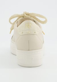 Baskets beiges avec des lacets texturés noués en nœud, languette en suède avec des clous dorés, et logo "pg" en relief sur la semelle et la languette.