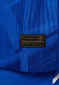 Tissu bleu texturé avec une étiquette noire et dorée portant l'inscription "ENGINEERED MB367590GL", cousue près de l'ourlet.