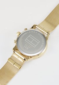 Goldfarbene Edelstahl-Uhr mit einem metallischen Mesh-Armband und rundem Ziffernblatt, graviert mit "TOMMY HILFIGER" und Wasserdichtigkeitsmarkierungen.