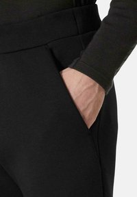 Pantalones negros hechos de una tela suave y elástica con un bolsillo lateral visible. La vista en primer plano muestra la cinturilla y una mano descansando en el bolsillo.