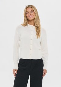 Usmievajúca sa žena s dlhými blond vlasmi, oblečená v bielom zapínanom kardigáne a čiernych nohaviciach s vysokým pásom na jednolatom pozadí.