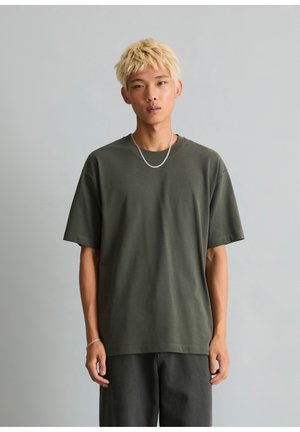 Jong persoon met kort blond haar, draag een donkergroen oversized T-shirt, zwarte broek, een kettingketting en een armband, staande tegen een grijze achtergrond.