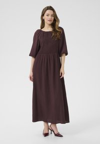 Longue robe bordeaux avec un buste smocké, des manches ballons et une jupe fluide. Le tissu présente un motif texturé en côtes. Assortie à des talons pointus.