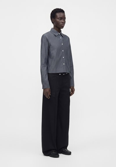 Personne portant une chemise grise à fines rayures boutonnée et un pantalon noir à jambes larges, debout devant un fond clair uni.