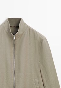 Veste beige clair avec col montant, fermeture éclair devant et poches latérales, en tissu texturé.