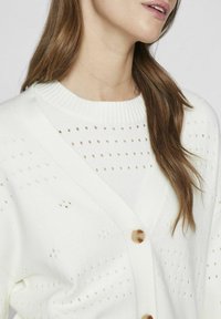 Bela kardigan z V-izrezom, gumbom na zapiranju in perforiranim vzorcem. Rebrast ovratnik in tekstura dodajata globino mehkem materialu.