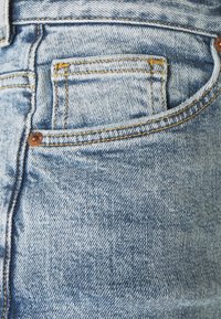 Jeans de mezclilla azul claro con un bolsillo trasero con costuras amarillas y un detalle de remache de cobre, mostrando una textura desgastada.