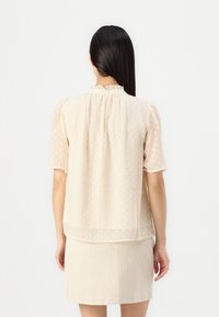 Kortermet, lys beige bluse med et strukturelt prikkemønster, rynket halslinning og løs passform. Kombinert med en beige skjørt.