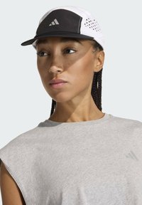 Cappello bianco e nero con perforazioni traspiranti, caratterizzato da un pannello frontale nero e un logo con tre strisce bianche. Indossato con un top grigio senza maniche.