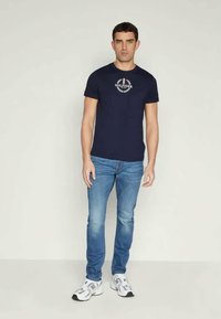 T-shirt de algodão azul marinho com mangas curtas e decote redondo, apresentando um logótipo em branco e vermelho. Combinada com jeans de tom azul claro e sapatilhas de corrida.